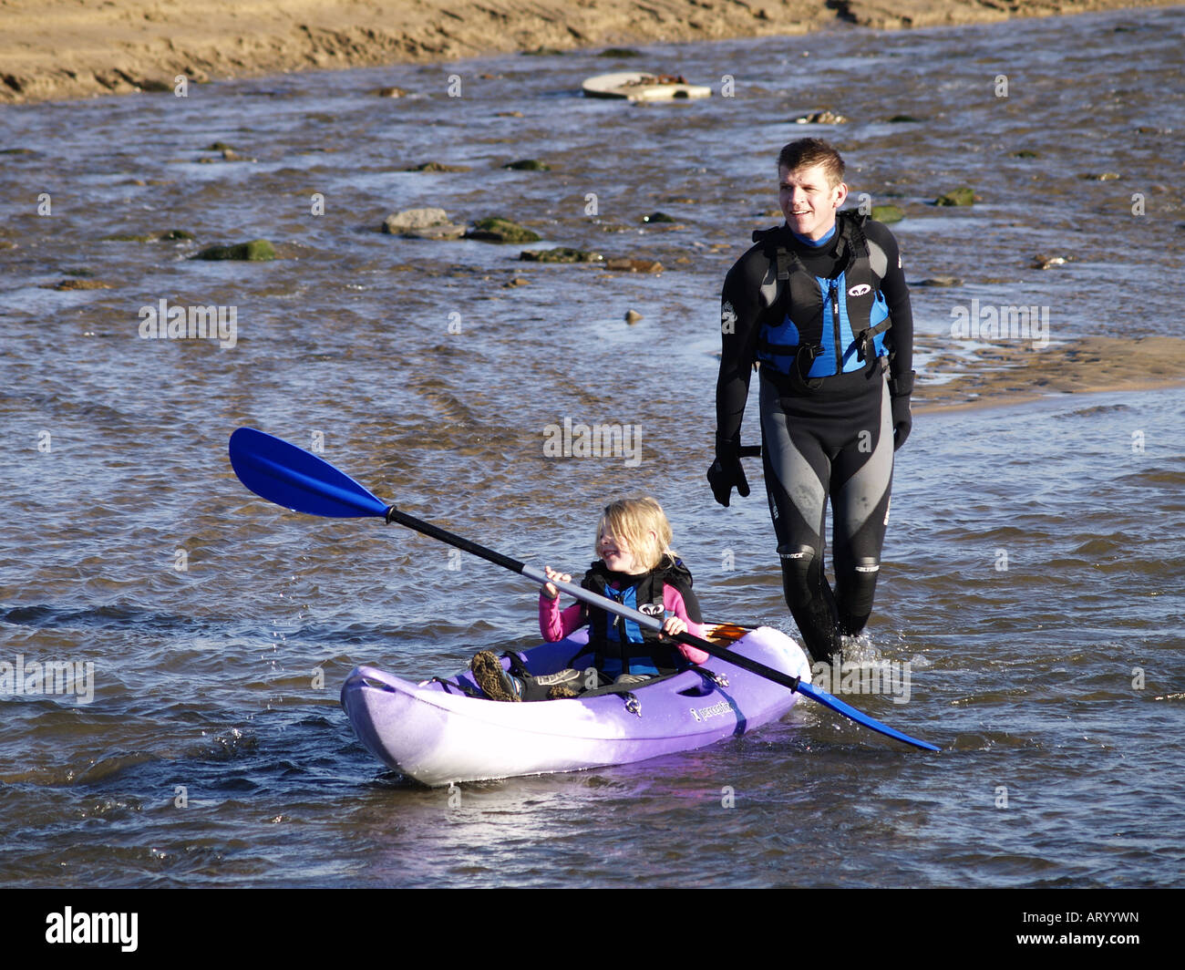 Young girl paddling surf ski canoe in shallow water at the beach, closely followed by dad Stock Photo