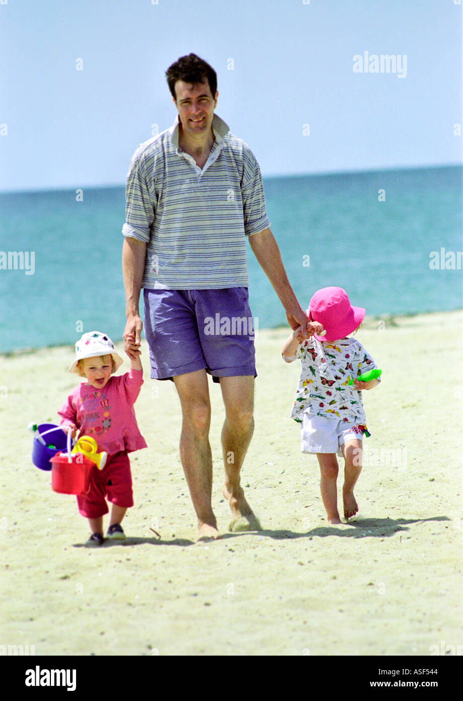 Father walking along the beach holding two young children with buckets and spades Stock Photo