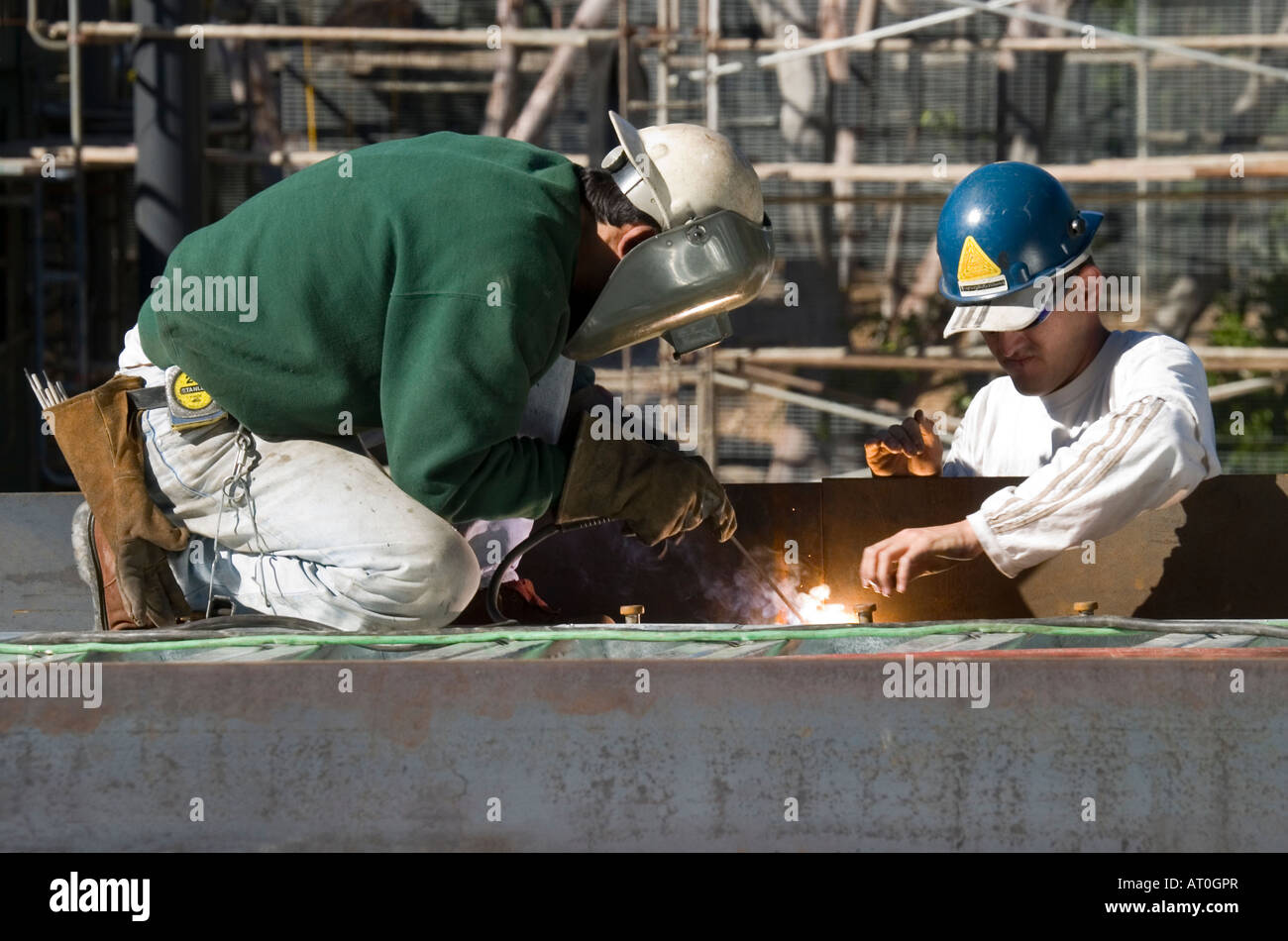 Two men in hard hats, working at a construction site.  One of the workers wears a protective face mask while spot welding. Stock Photo