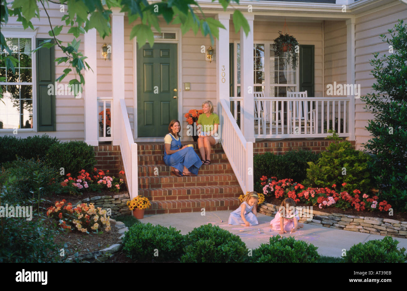 Two mothers visit on the steps of a front porch while their children play in a suburban neighborhood Stock Photo
