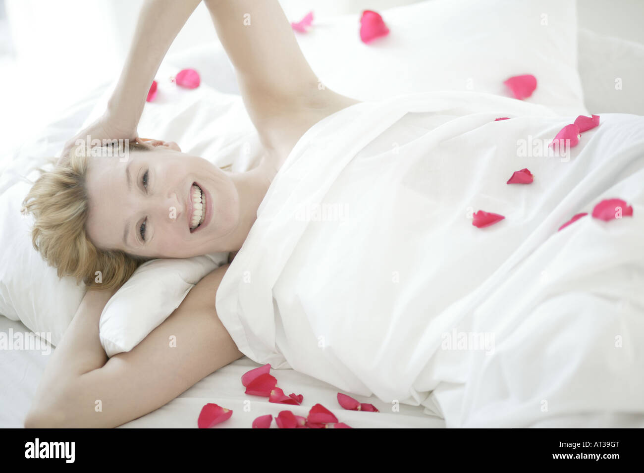 A woman lying in bed smiling, rose petals scattered on  the bed Stock Photo