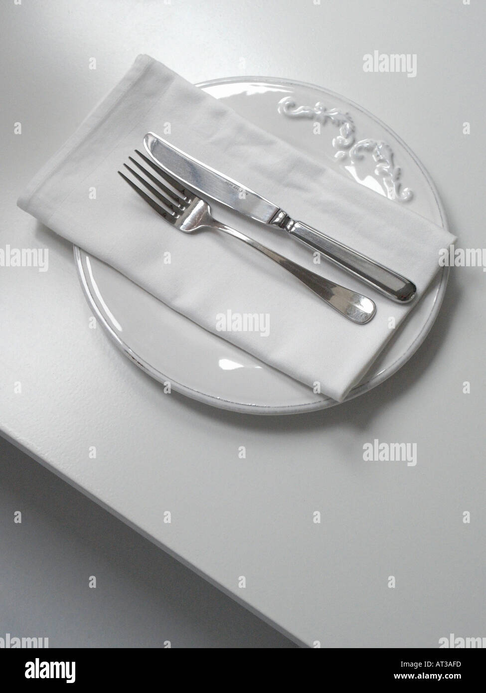 Vertical shot of a white simply decorated plate with napkin silver knife and fork on a white table Stock Photo