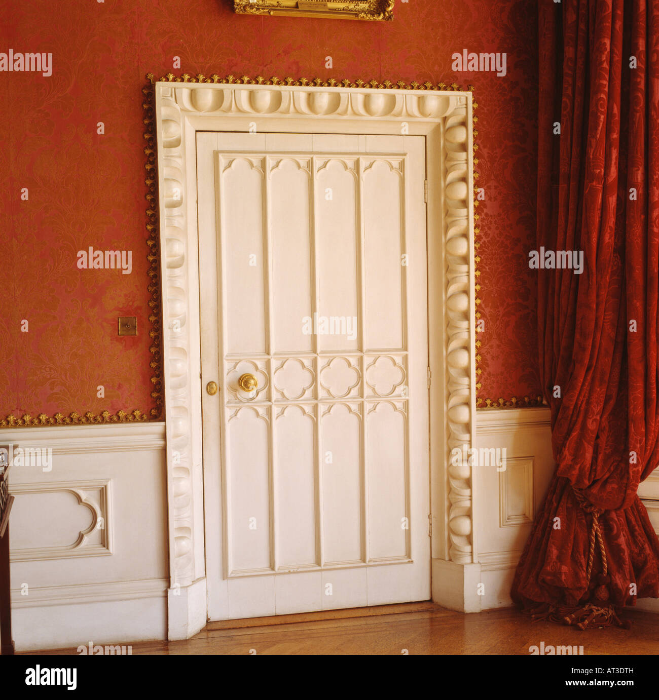 Close up of white Gothic door with ornately carved frame in red room with white wainscot paneling and red silk curtain Stock Photo