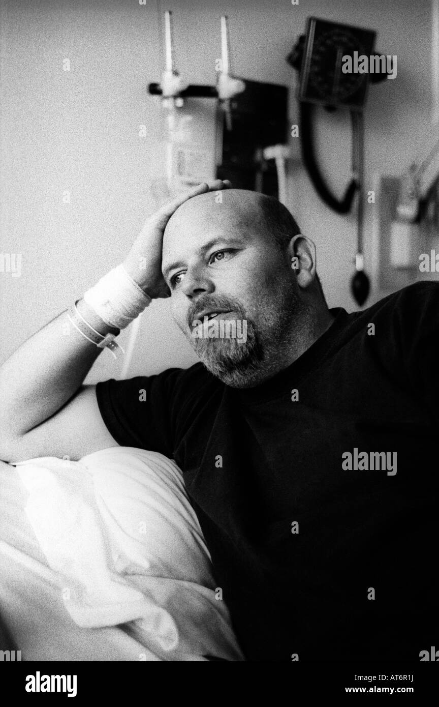 Large balding bearded man in hospital bed rests head on hand and gazes