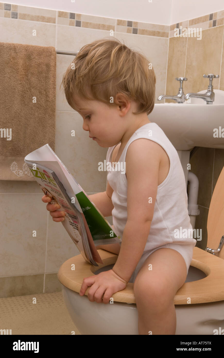 boy toddler two years old, sitting on toilet, loo, reading magazine, wearing a white singlet vest Stock Photo