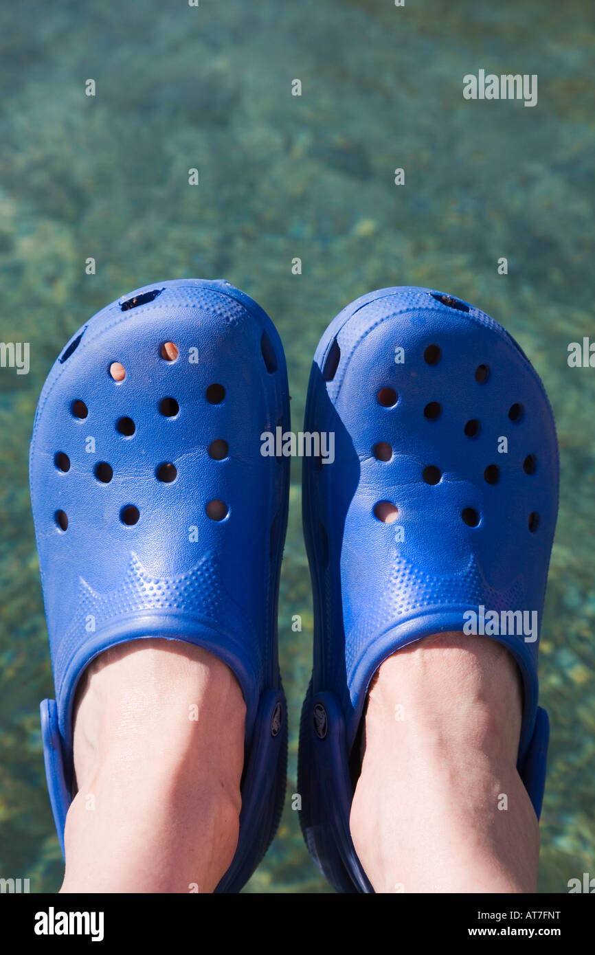 Pair of person's feet wearing blue Crocs plastic sandals against clear sea water Stock Photo