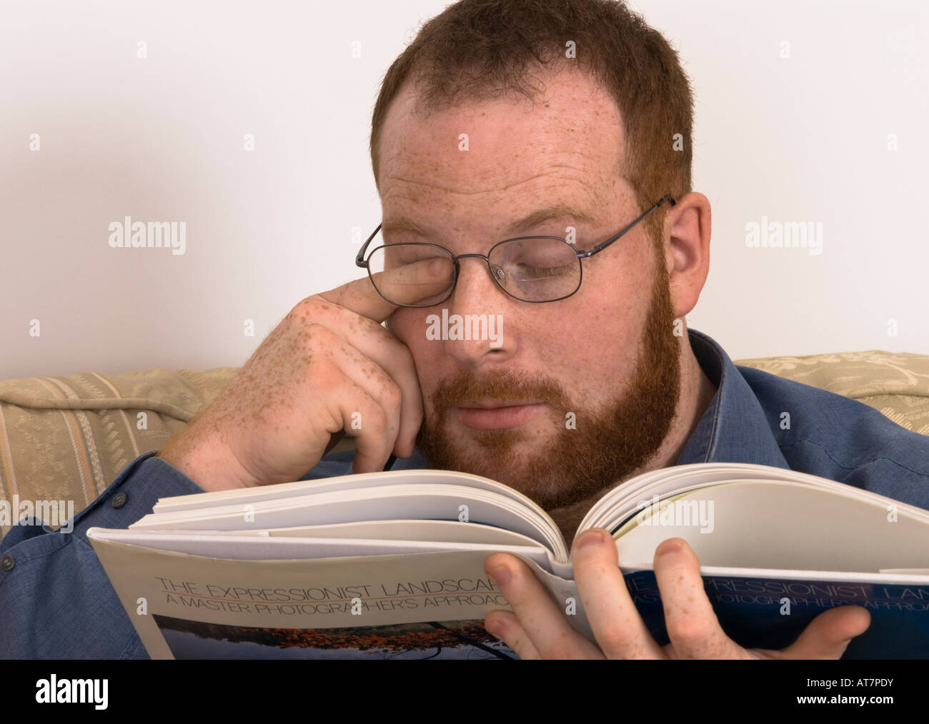 young man with poor eyesight wearing glasses rubbing his strained eye with finger trying to read book, incorrect spectacles Stock Photo