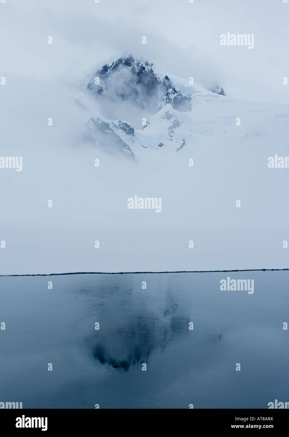 Peak of an antarctic mountain showing through the mist and reflected in the deep blue waters beneath Stock Photo