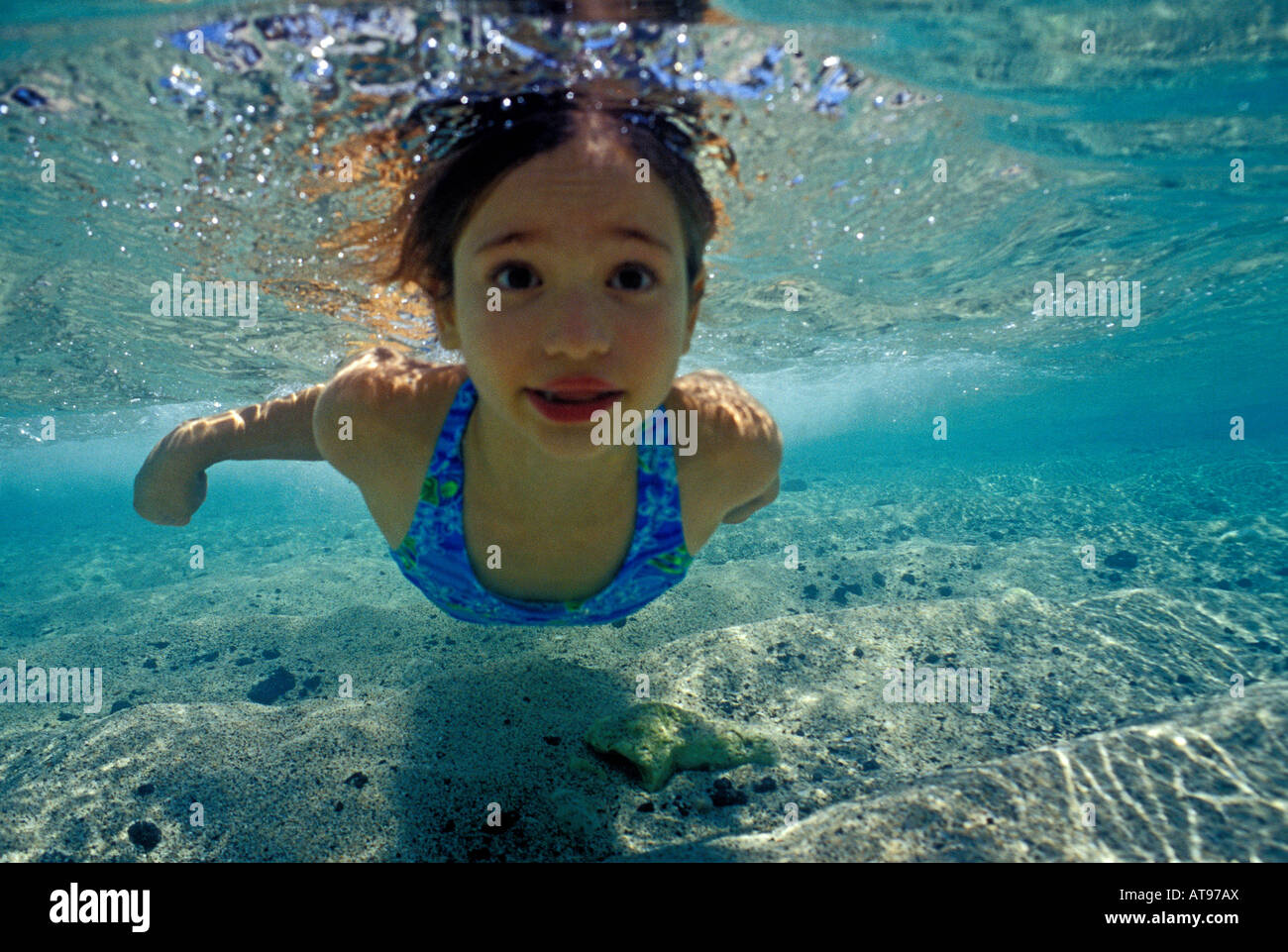 A young girl with eyes open swims underwater in the clear, blue shallow