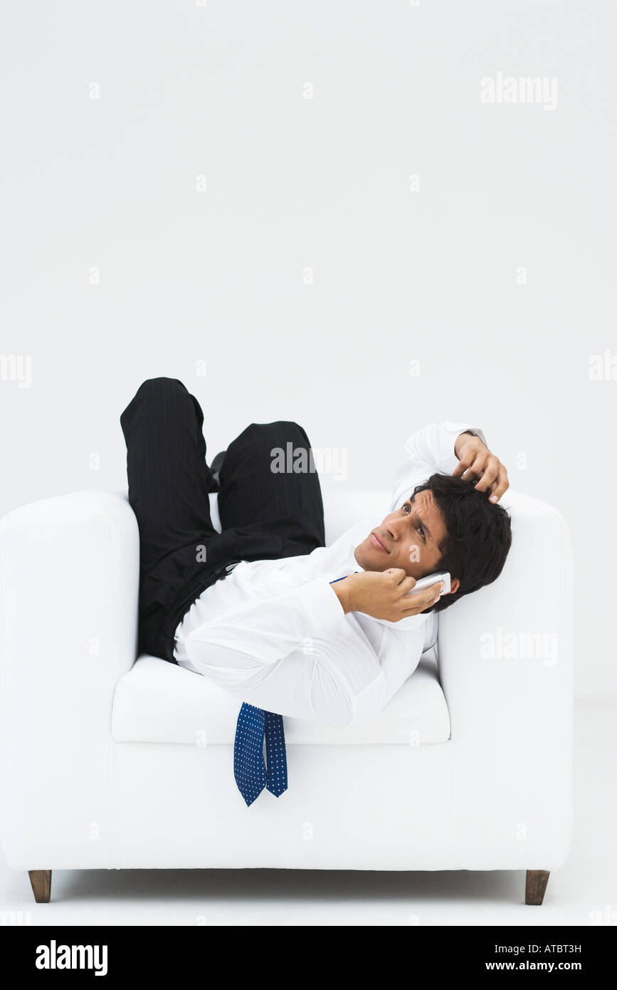Businessman lying backwards in armchair, holding cell phone to ear and scratching head, full length Stock Photo
