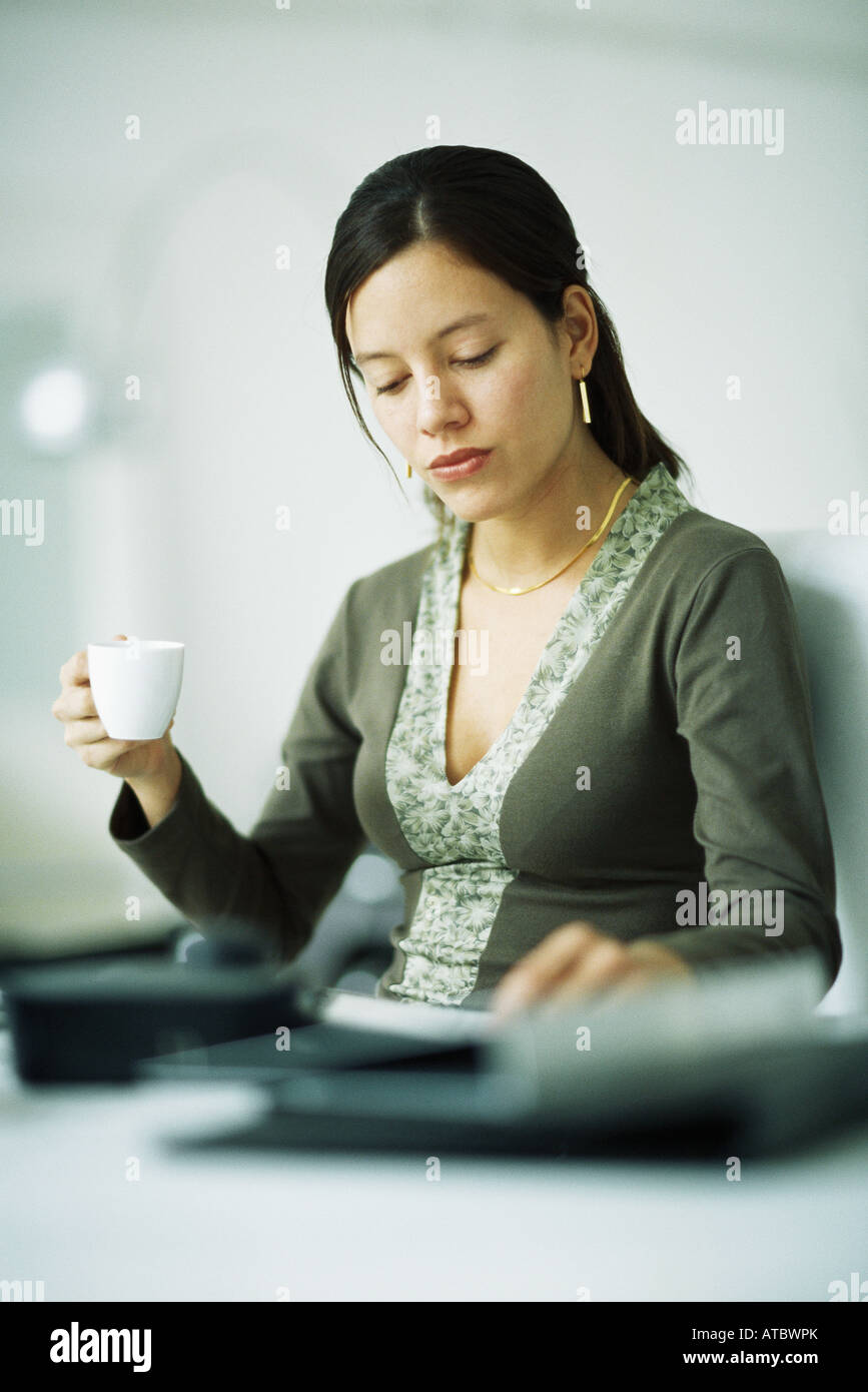 Woman sitting at desk, holding coffee cup, looking down Stock Photo