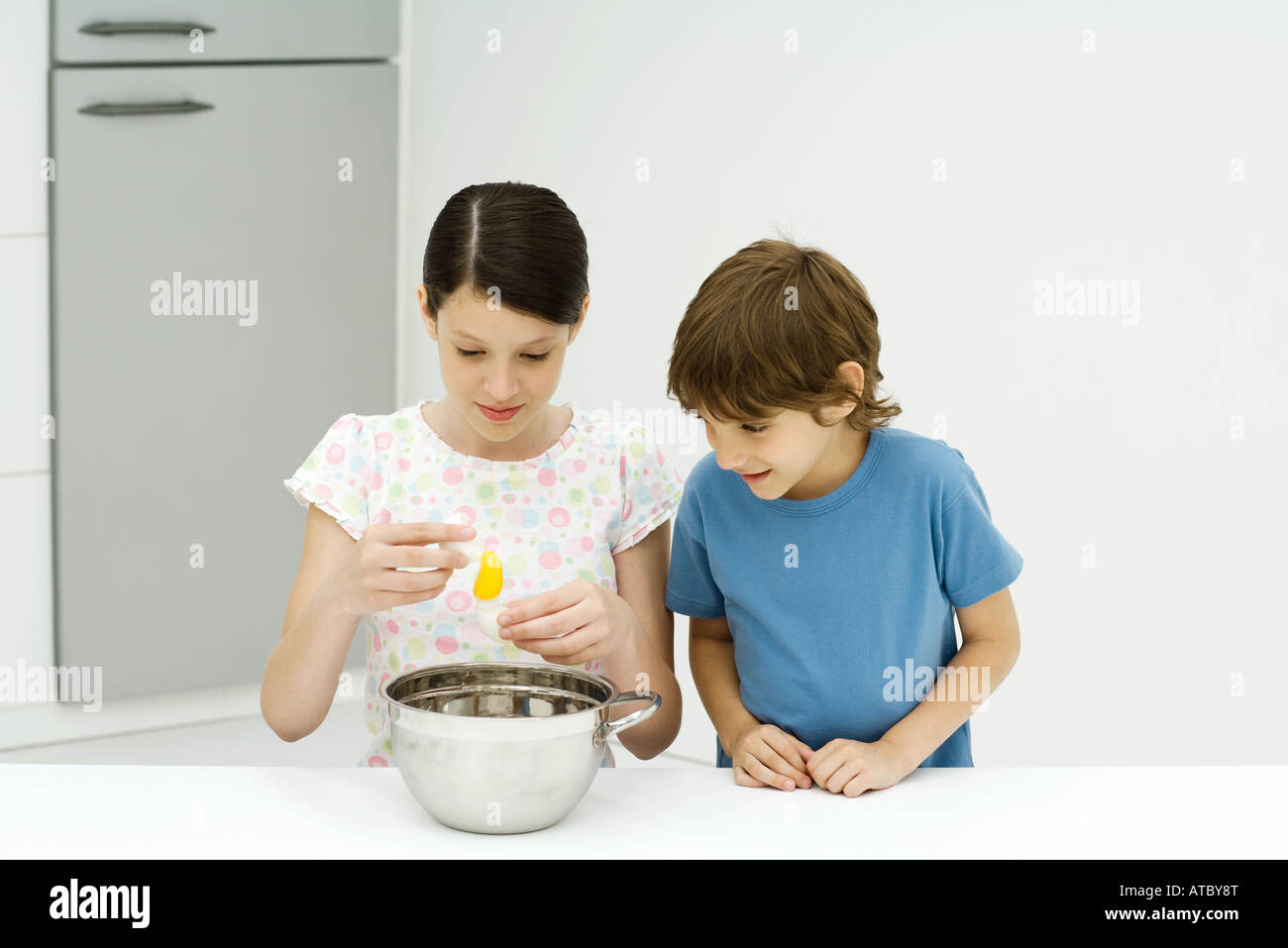 Two young siblings cooking together, girl cracking egg, both looking ...