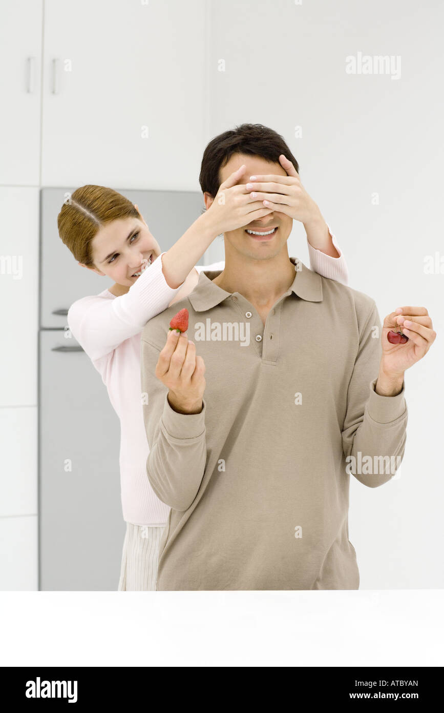 Woman standing behind man with her hands over his eyes, man holding strawberry in one hand, cherries in the other Stock Photo