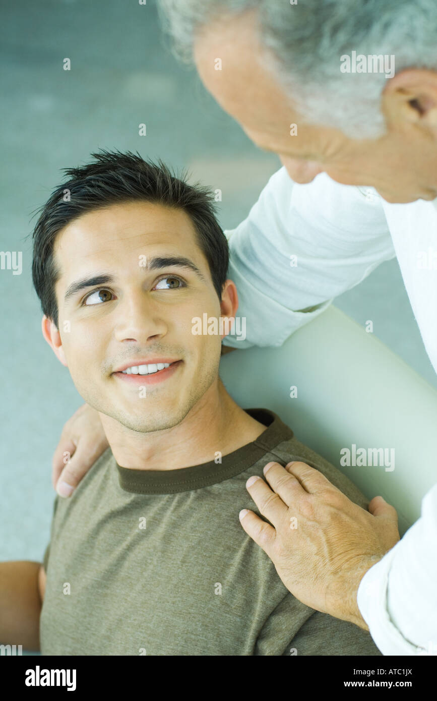 Mature man leaning over, placing hands on son's shoulders, both smiling at each other Stock Photo