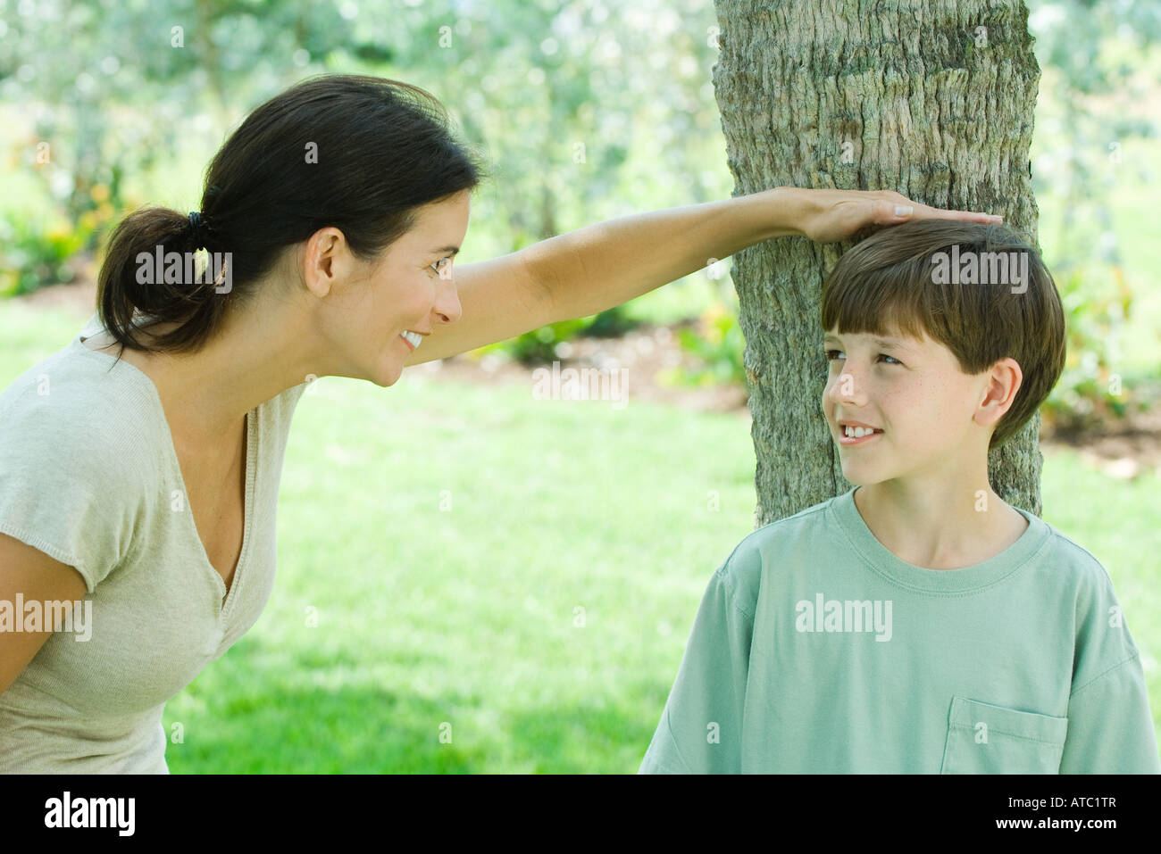 Boy leaning against tree trunk, his mother placing her hand on his head, both smiling Stock Photo