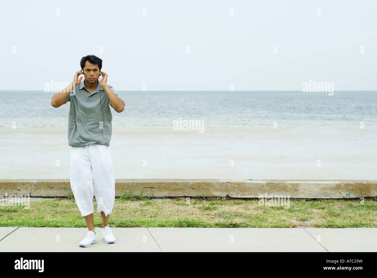 Man standing in front of beach, listening to headphones, hands raised to head Stock Photo