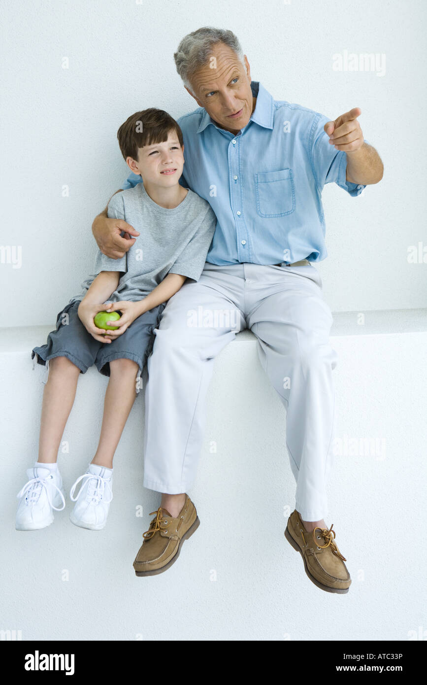 Man sitting with arm around grandson's shoulders, pointing, both looking away Stock Photo