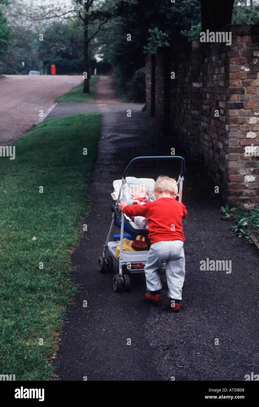 Little boy of about three 3 years old in a red top pushing his baby sister along a pavement sidewalk in a pram Stock Photo