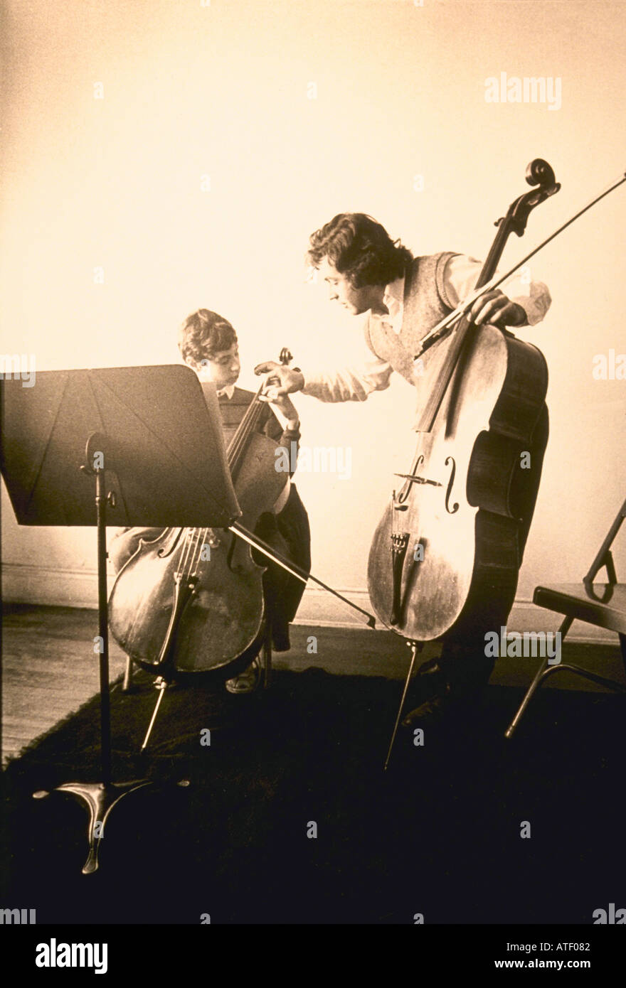 Hand colored image of music teacher with cello bending over to fix 12 yo male student hand as he sits with his cello Stock Photo