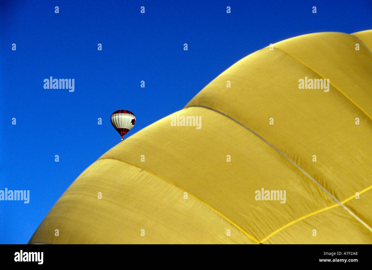 Yellow balloon being filled with hot air ready for flight with smaller balloon already in flight in distance. Stock Photo
