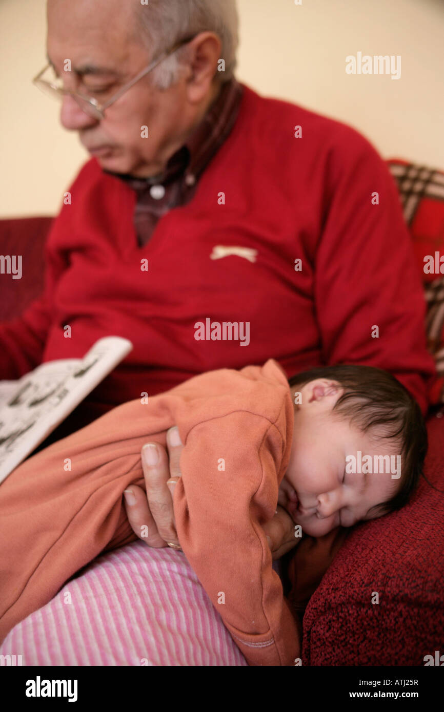 One week old baby sleeps on grandad's lap while he reads the newspaper Stock Photo