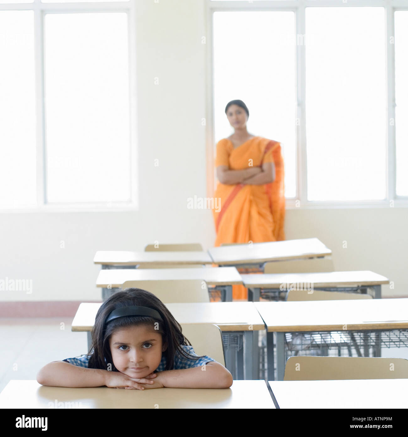 Portrait of a schoolgirl leaning on a desk with a teacher standing in ...