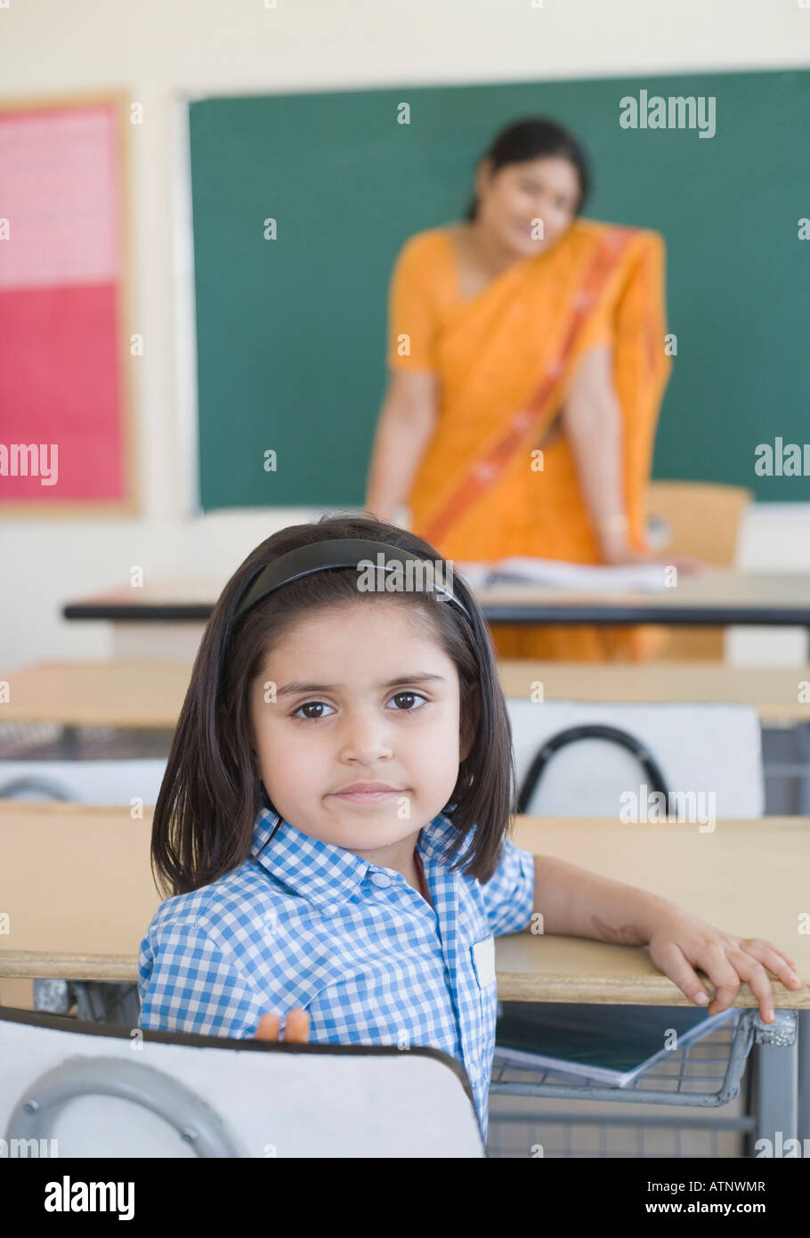 Portrait of a schoolgirl sitting at a desk with her teacher standing in ...