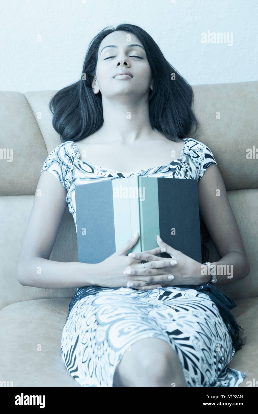Young woman sitting on a couch with her eyes closed and holding a book ...