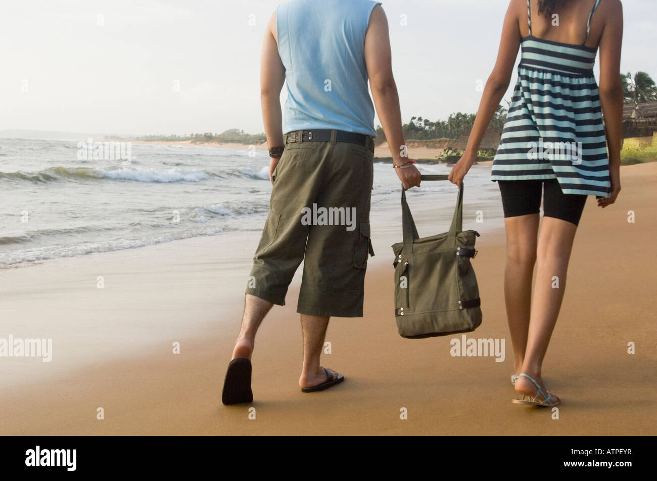 Rear view of a young couple holding a bag and walking on the beach ...