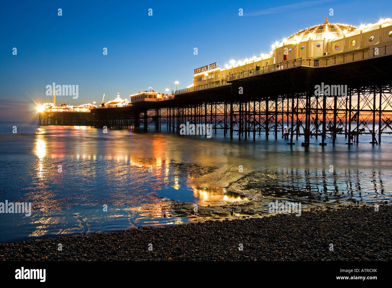 Brighton Pier in low light at sunset with colourful lights on pier reflecting in the sea and pebble beach in foreground Stock Photo