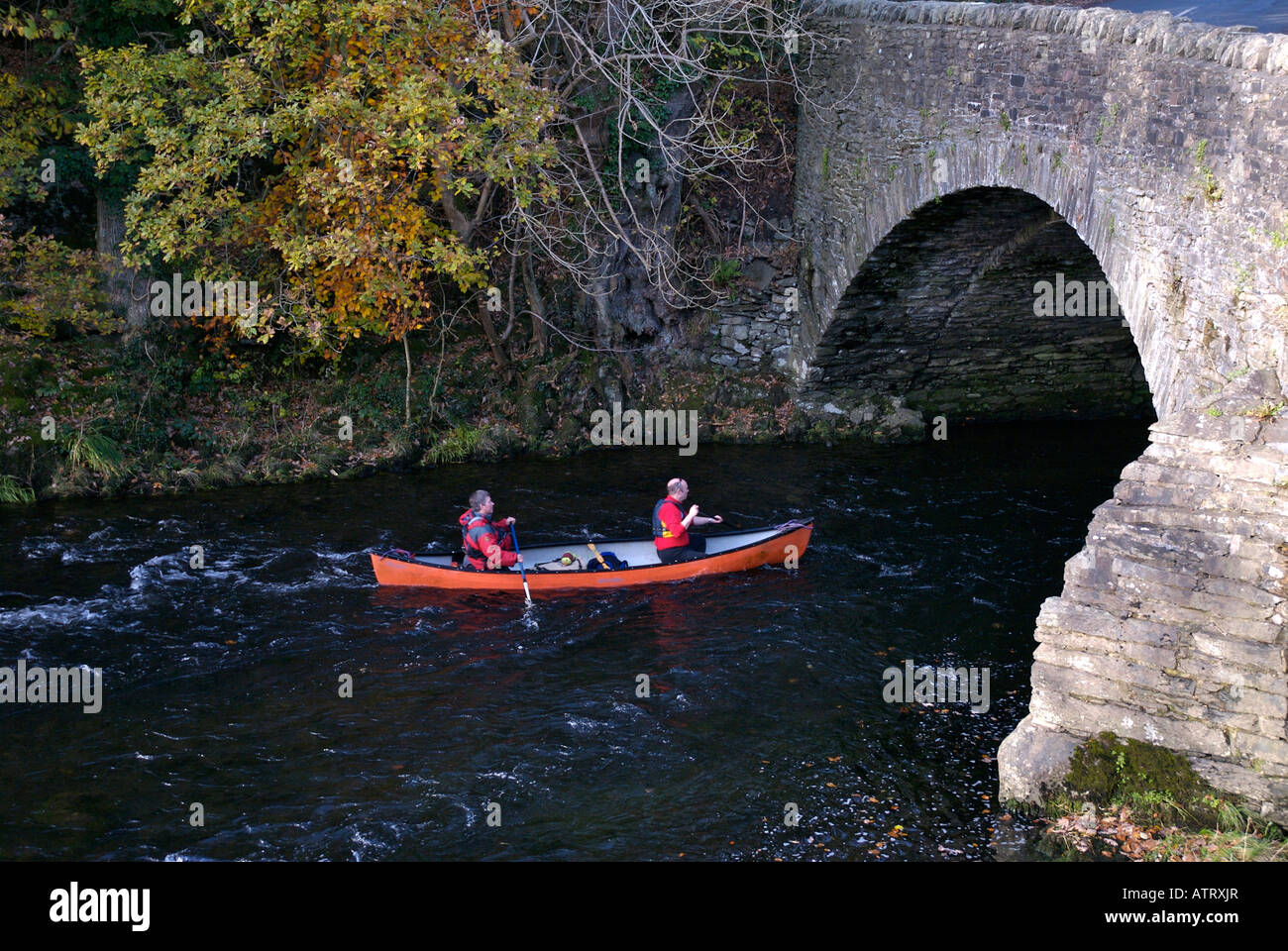Two people paddling open canoe under a bridge, River Brathay at