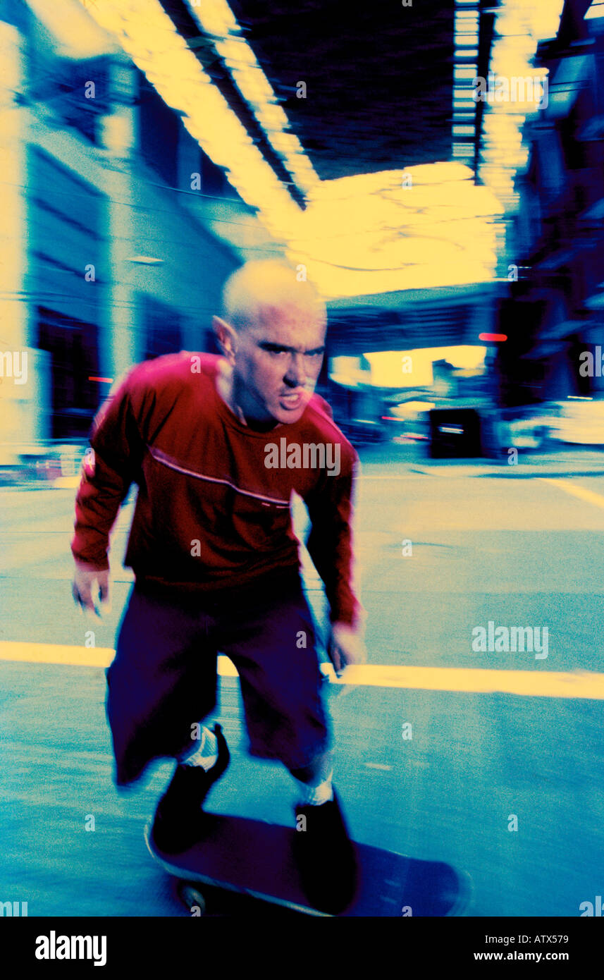 A skateboarder with a shaved head displays a hostile attitude as he traverses a city street Seattle Washington Stock Photo