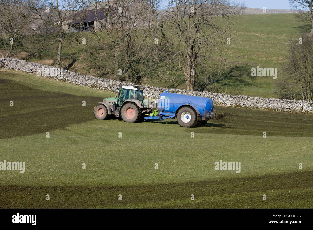 Farmer working driving green tractor & cylindrical fertilizer tank spraying discharging slurry on farmland pasture field grass - Yorkshire, England UK Stock Photo