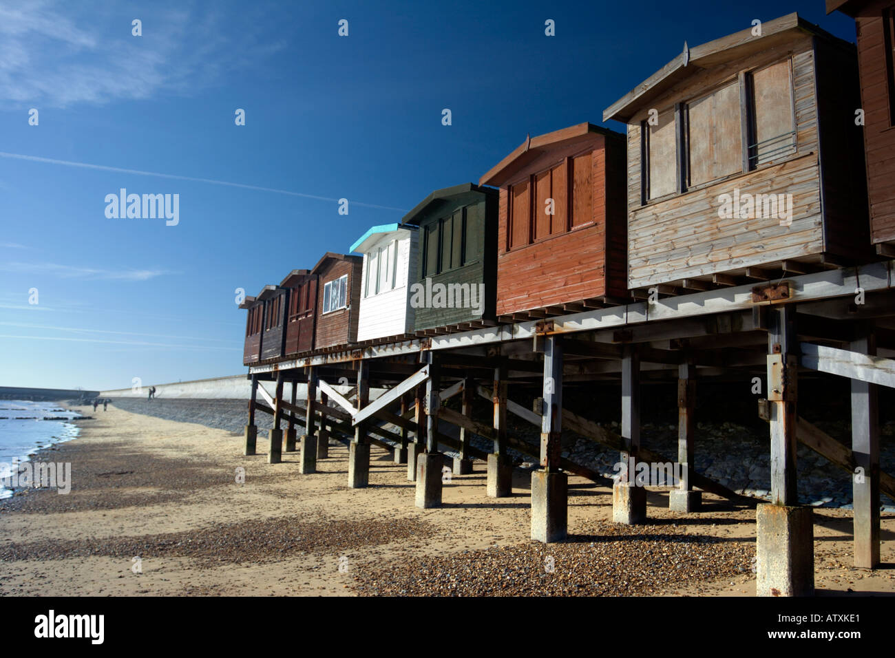 A row of traditional beach huts on stilts at Frinton on sea, showing the concrete sea wall behind Stock Photo