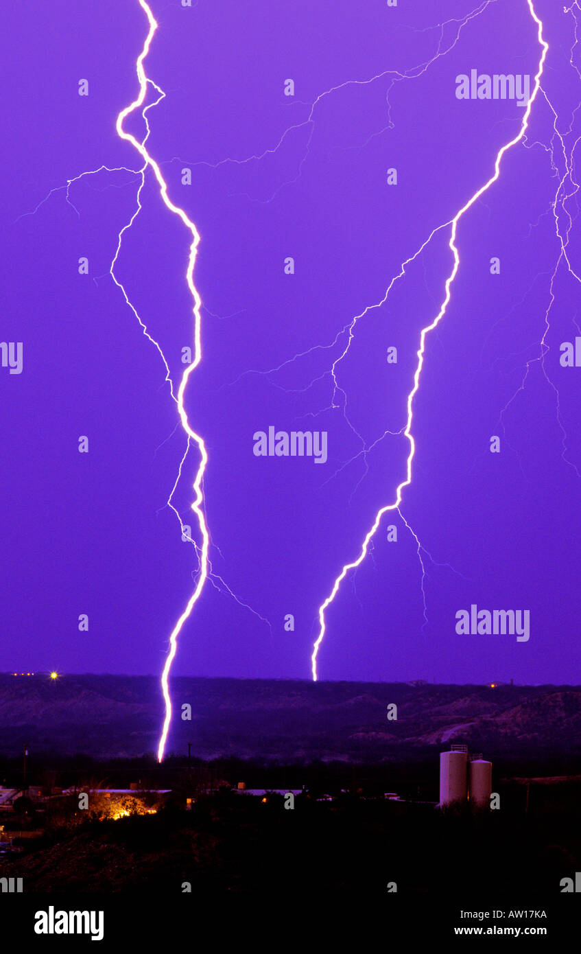 Lightning bolts strike down against a blue and purple sky with water towers and glowing lights from a town in Southern Arizona. Stock Photo
