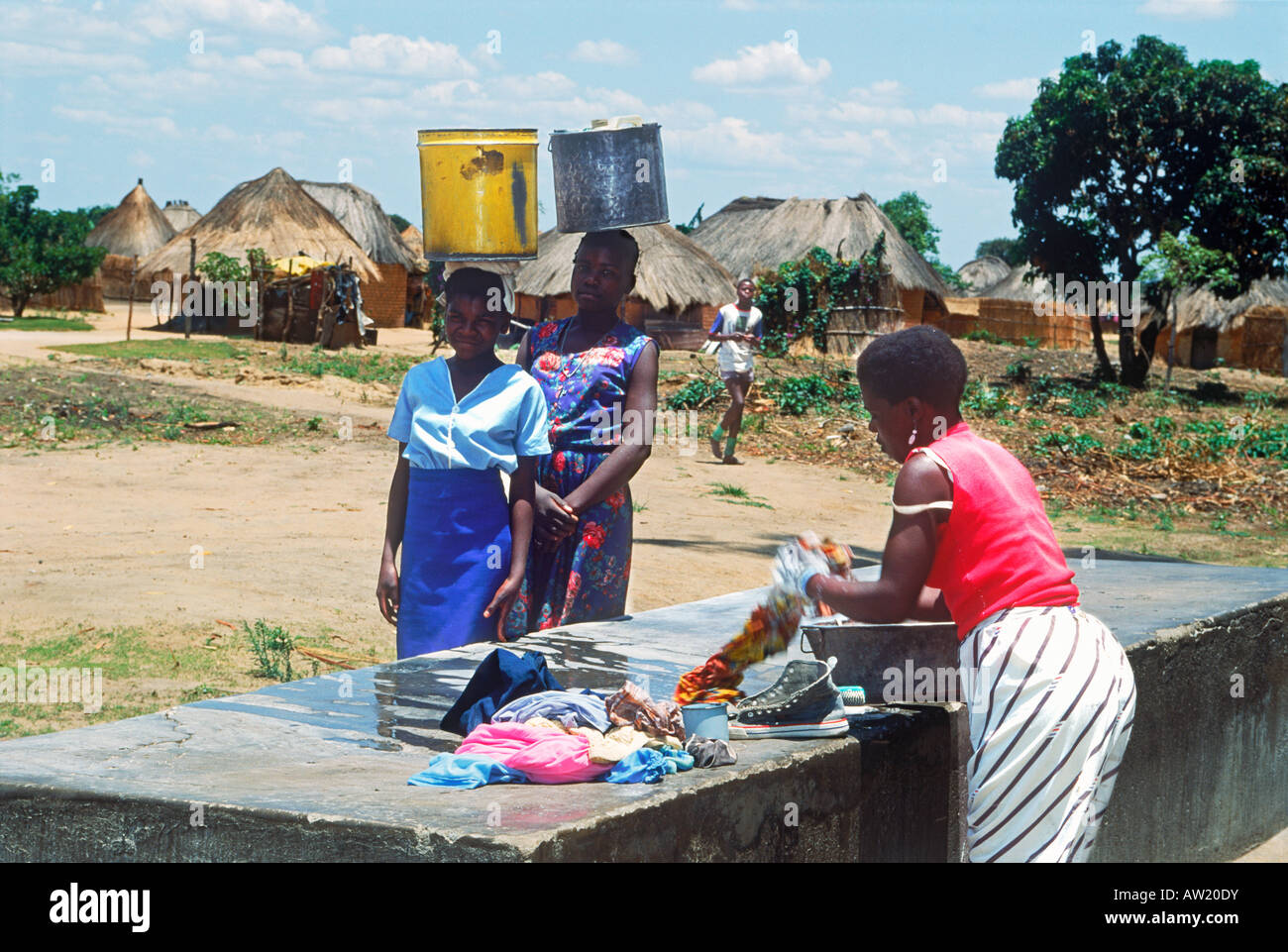 Young girls porting heavy buckets of water past woman washing clothes