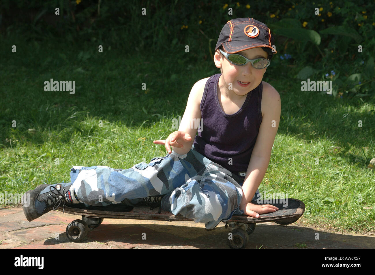 5 year old caucasian boy sitting on a skateboard wearing baseball hat and sunglasses Stock Photo