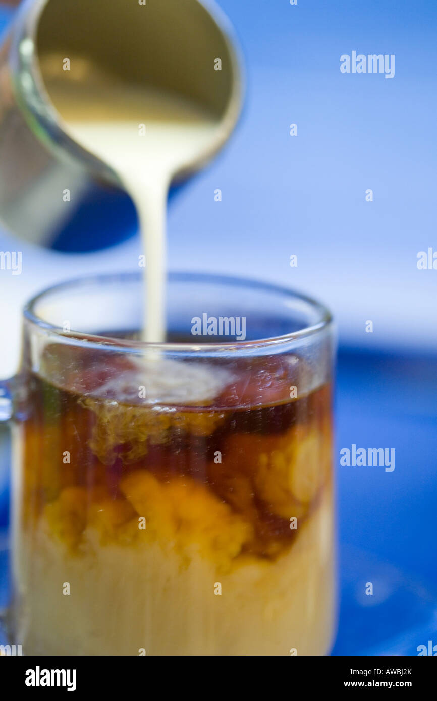 Sequence of pouring milk from a stainless steel jug into a glass of tea against blue background Stock Photo