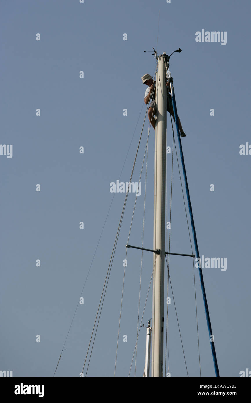 A man works at the top of the mast of his sailboat making repairs and inspecting the rigging and instruments Stock Photo