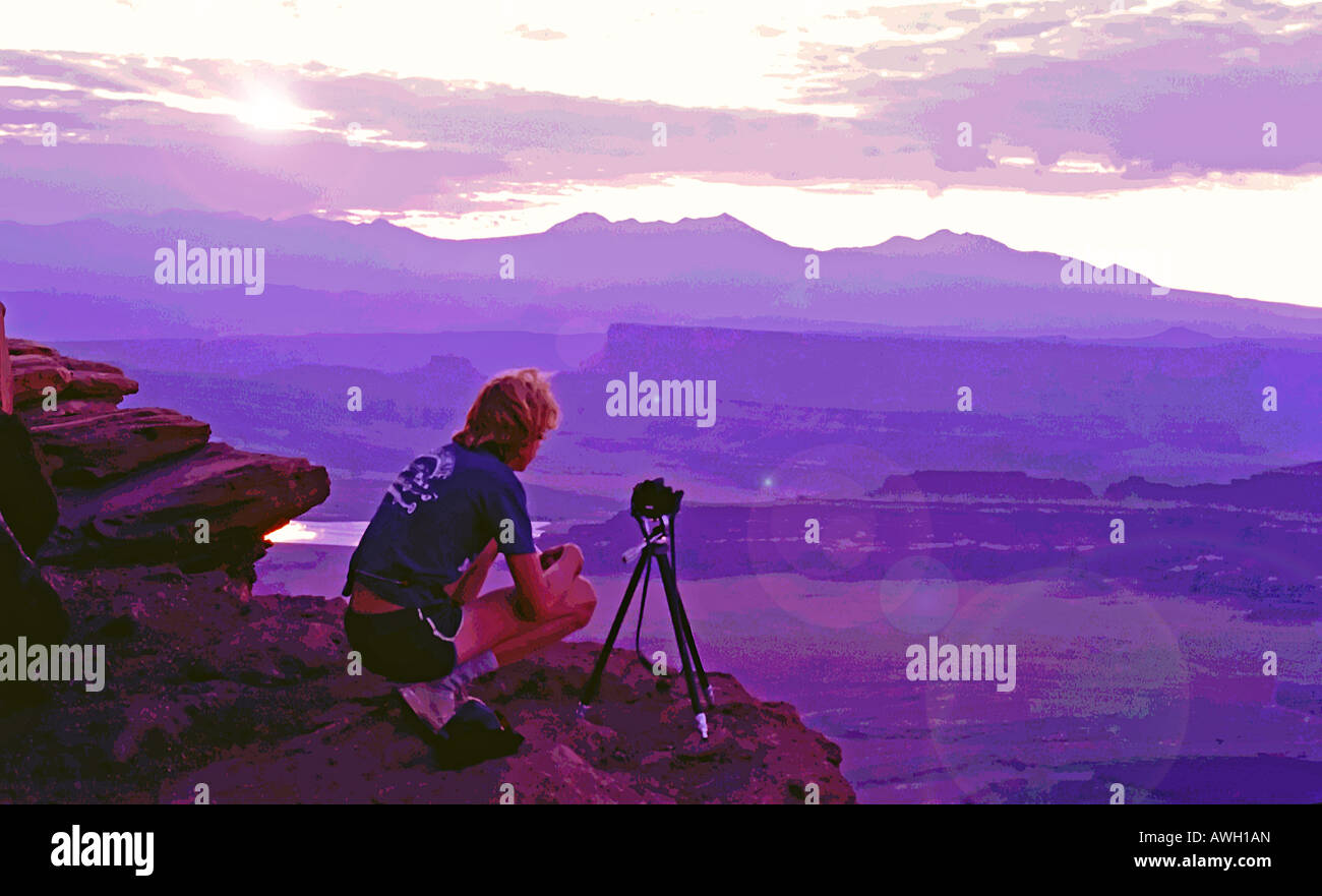 A young man perches near the edge of Dead Horse Point, Utah, USA with his tripod mounted camera at the ready as the sun rises. Stock Photo