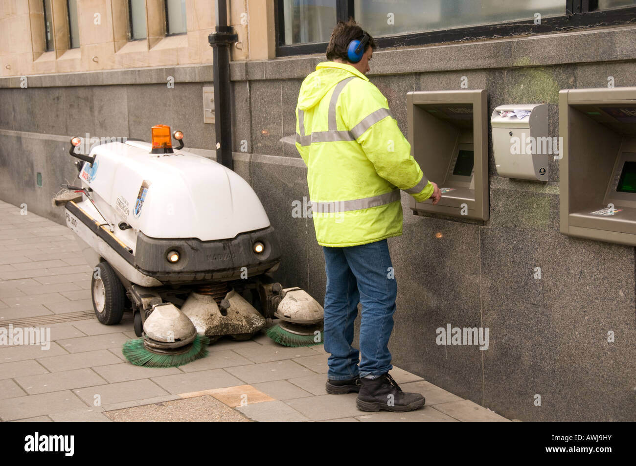 Local authority roadsweeper taking money out of ATM cashpoint machine with his vacuum cleaner machine by his side Stock Photo