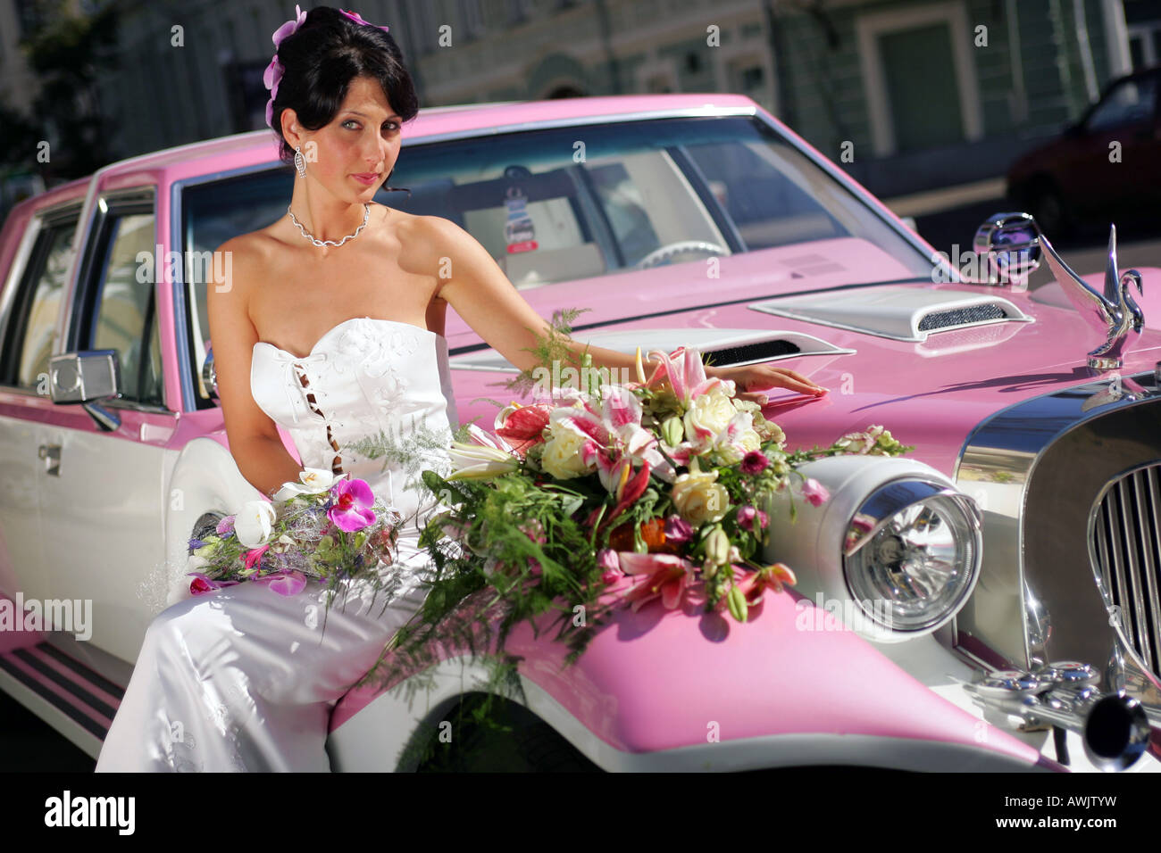 Beautiful smiling bride in white dress on wedding day. She is sat on the hood on a pink cadillac limousine wedding car Stock Photo