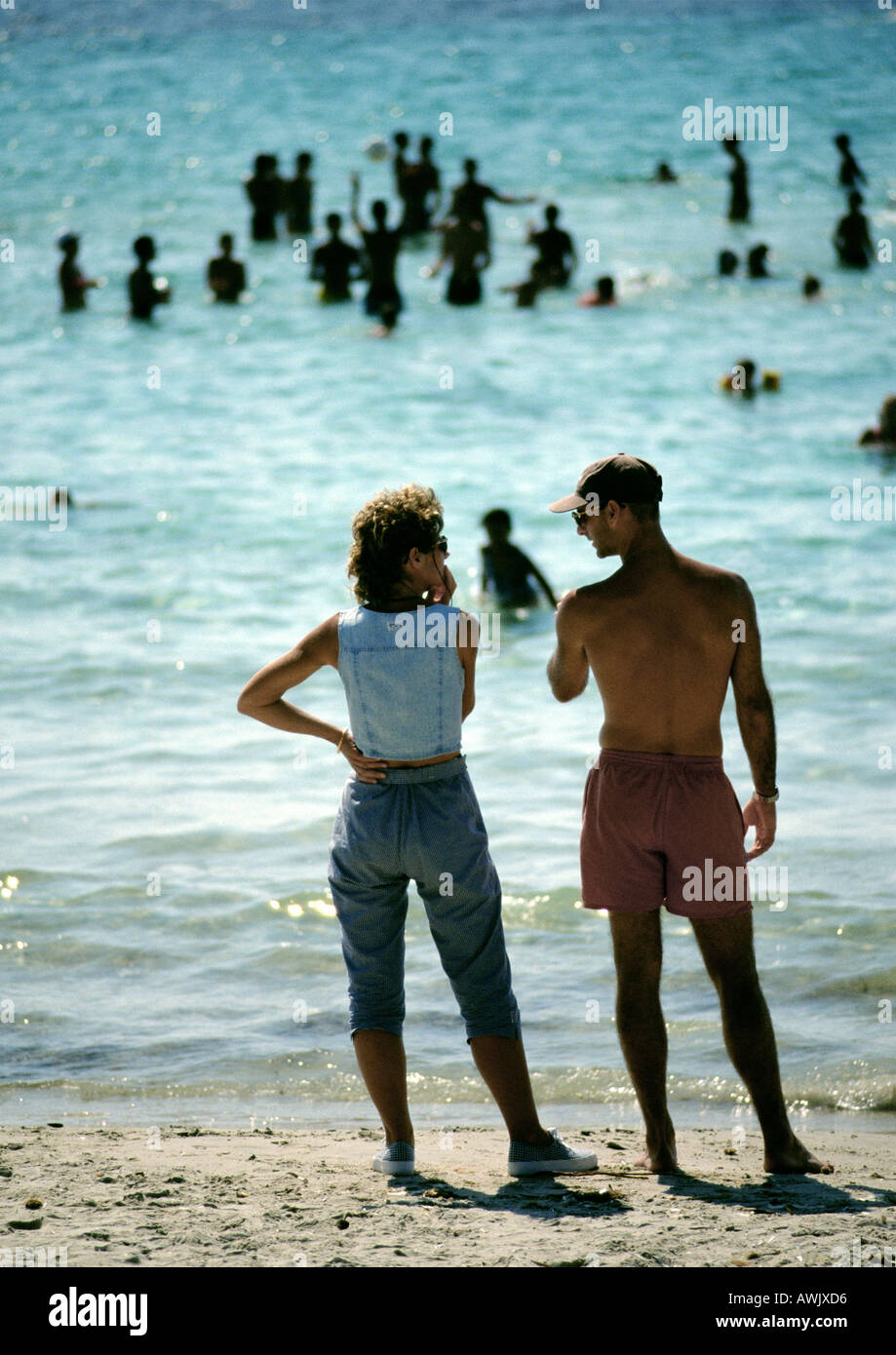 People standing at the beach, people playing in the water in background, rear view. Stock Photo