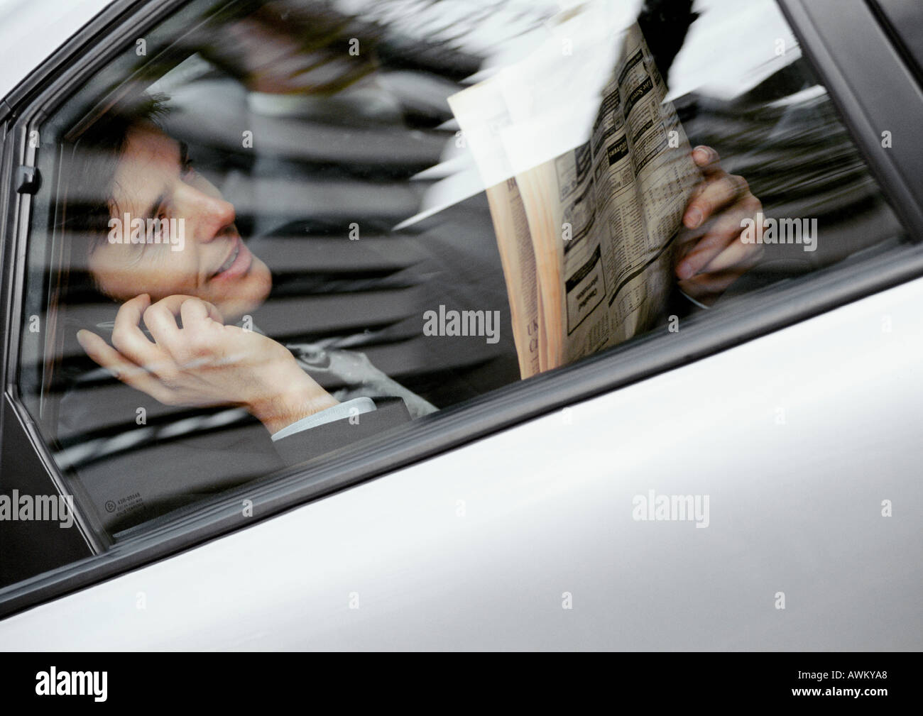 Businessman sitting in back of car, holding newspaper and cell phone, viewed through window Stock Photo