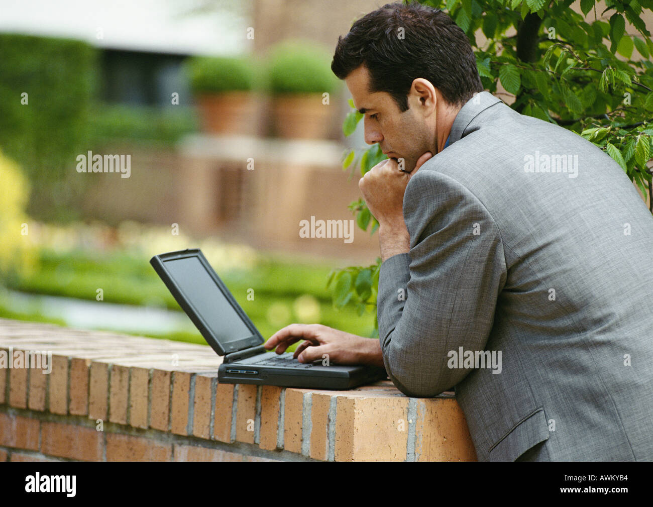 Businessman using laptop computer outside, leaning against low brick wall, side view Stock Photo