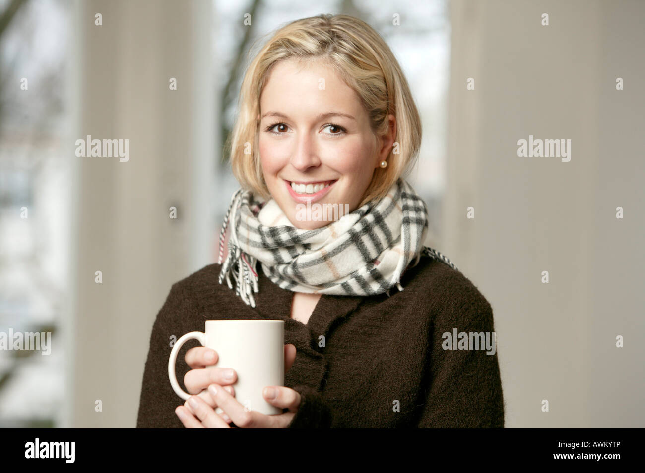 Smiling young blonde woman in a dark sweater and wool scarf holding a mug Stock Photo