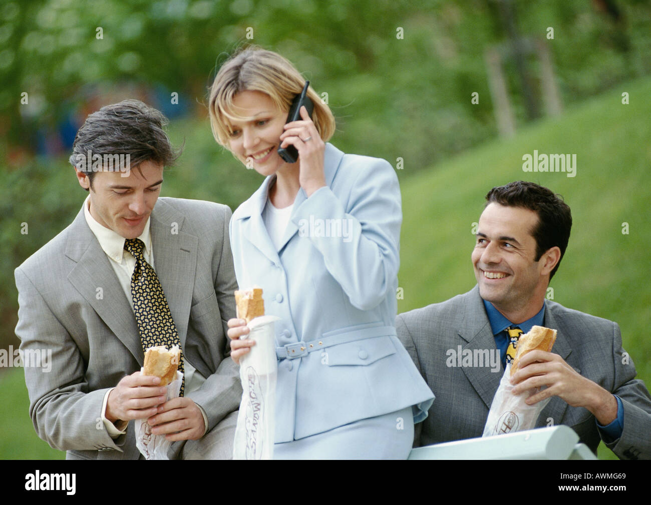 Three business people holding sandwiches outdoors, one using cell phone Stock Photo