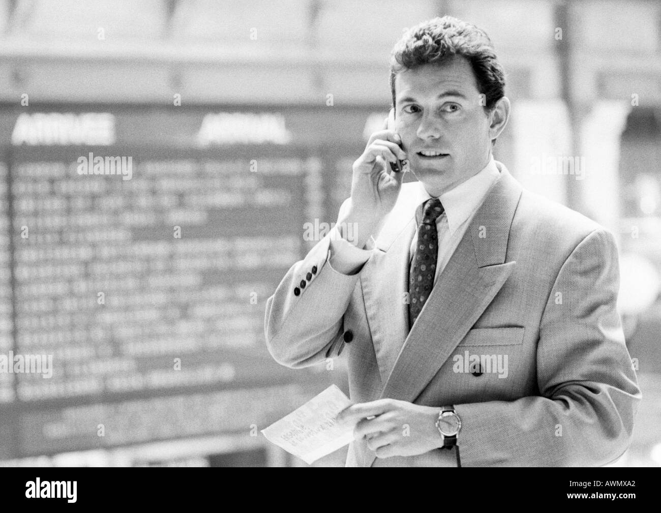 Businessman using cellular phone, standing in front of arrival board, black and white. Stock Photo