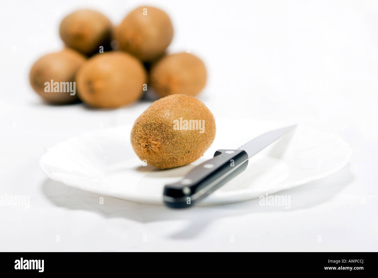 Single kiwi on a plate with knife, more kiwis piled at the back Stock Photo