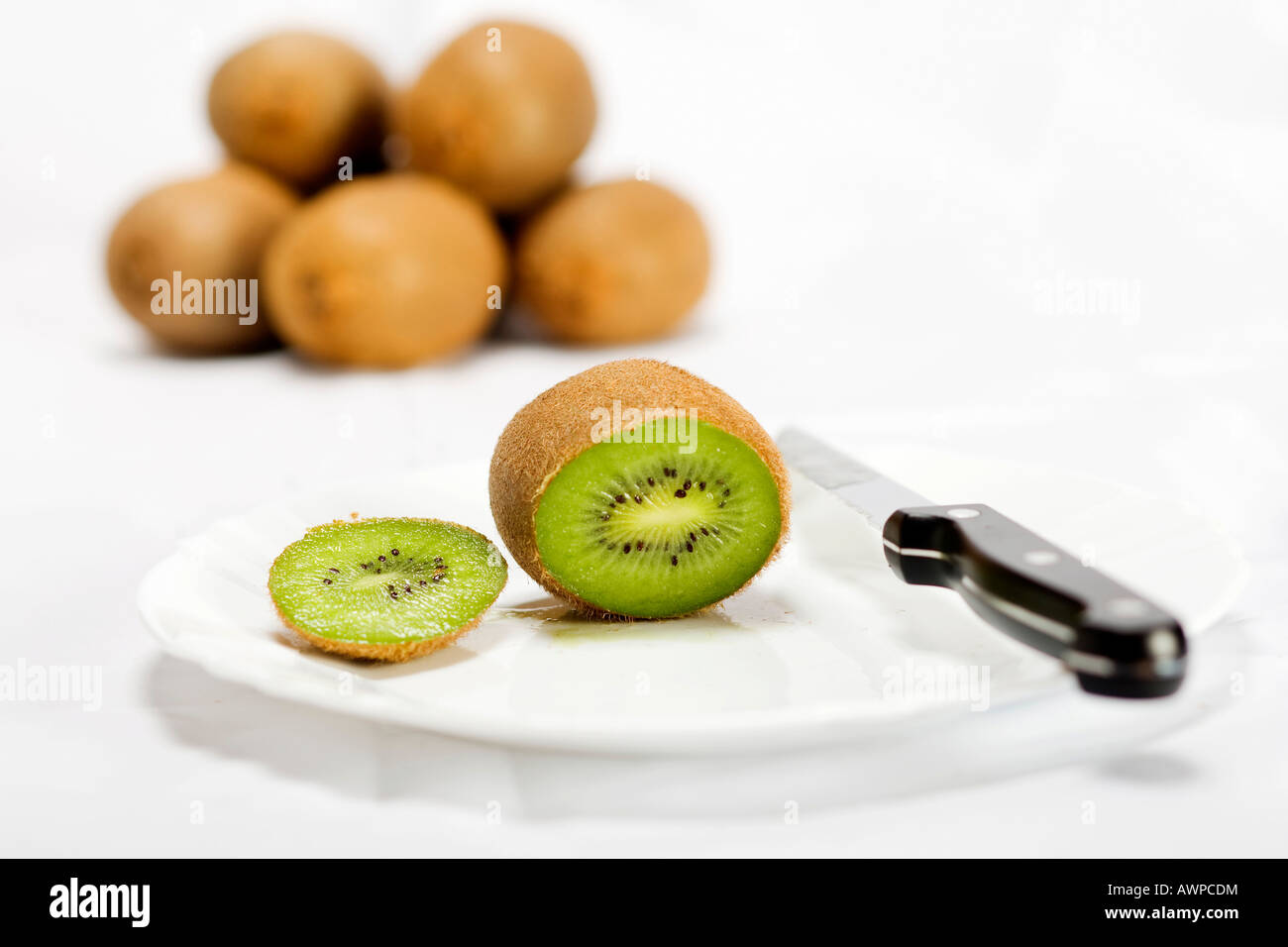 Single kiwi cut open on a plate with knife, more kiwis piled at the back Stock Photo